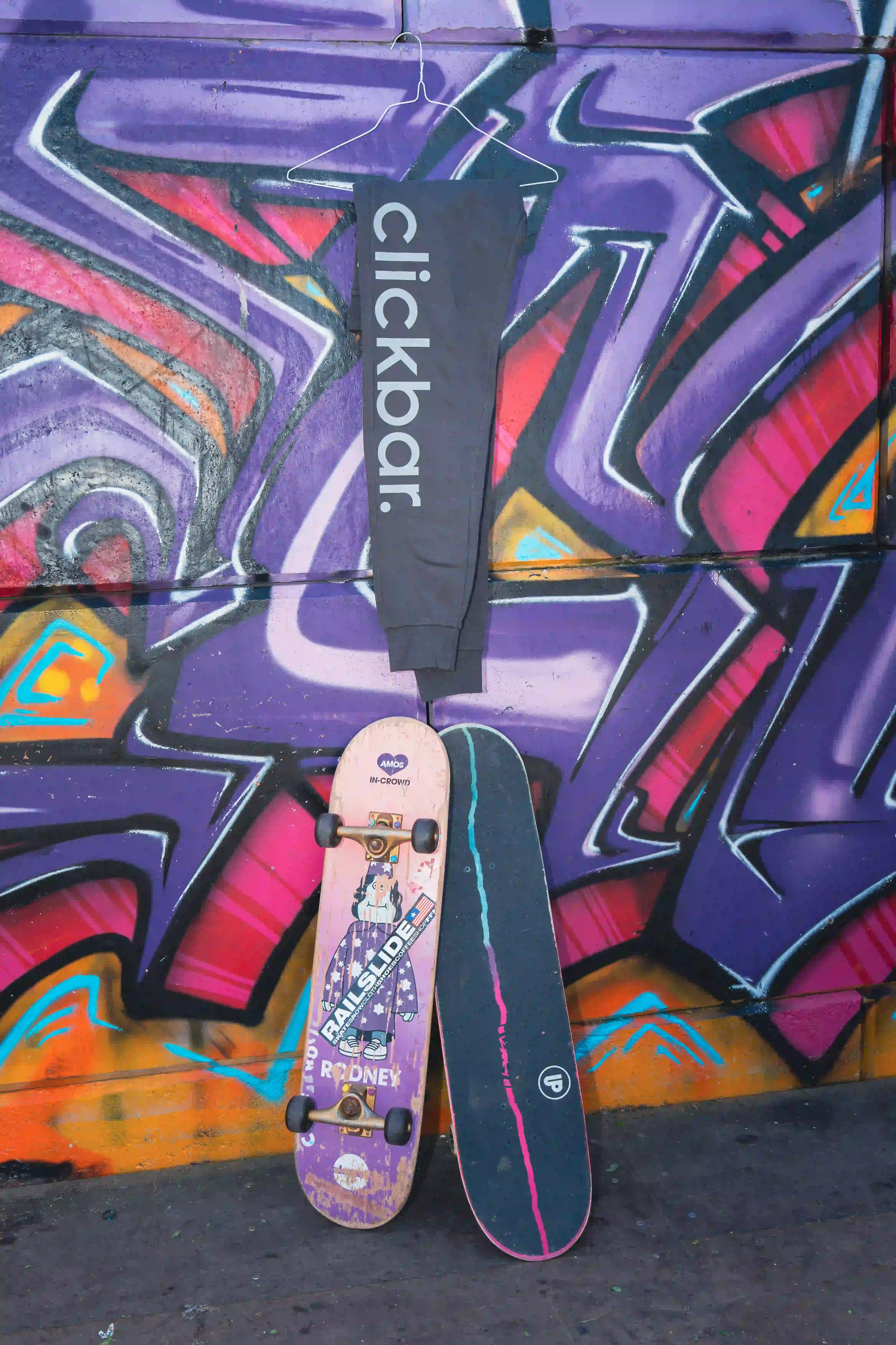 'clickbar.' Jogginghose an Kleiderbügel vor Graffiti-Wand, zwei Skateboards davor.